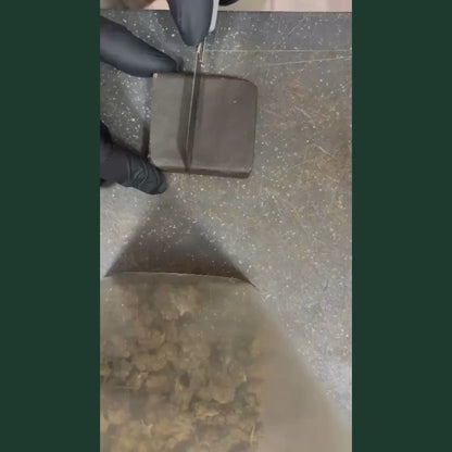 Video produit resine CBD Charras CSA2 : mains gantees de gants noirs manipulant un bloc de resine CBD compresse brun fonce. La video montre le produit sous plusieurs angles, puis le bloc brise en deux revelant un interieur texture doree et granuleuse. Fond vert, surface granuleuse au sol. Resine CBD artisanale haute concentration, legale moins de 0,3% THC, disponible a l achat en France.