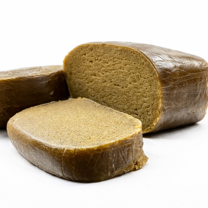 Deux morceaux de resine CBD CSA 30% Piatella en tranche epaisse sur fond blanc uni. Bloc arriere-plan entier avec croute exterieure lisse brun fonce craquelée, reflets cireux. Tranche au premier plan revele l'interieur beige dore clair, texture granuleuse dense homogene et compacte, coupe franche nette. Contraste marque entre croute sombre et chair interieure claire. Concentre hash premium THC legal 0,3%, CBD Pas Cher 14, livraison France.