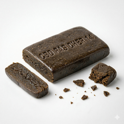 Bloc de resine CBD Charras CSA noire brillante de forme rectangulaire, inscription en relief CBD PAS CHER 14 sur la face superieure, texture grasse et collante, surface lisse avec reflets brun fonce. Au premier plan a gauche un barreau coupe, a droite un morceau detache accompagne de miettes eparpillees sur fond blanc uni. Hash premium notes terreuses boisees epicees, THC legal 0,3%, CBD Pas Cher 14.